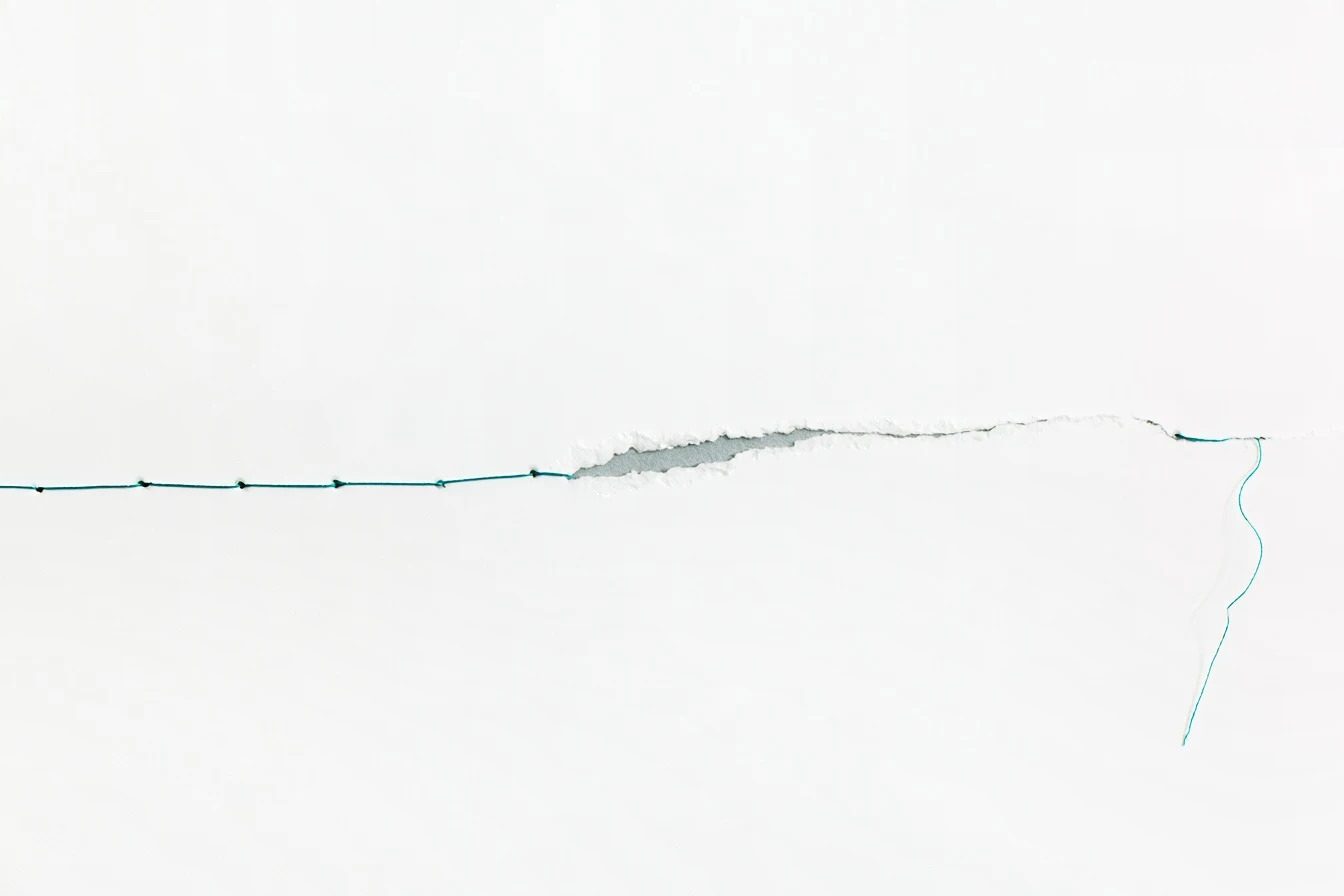A small tear in white paper, closed at one end with teal running stitch